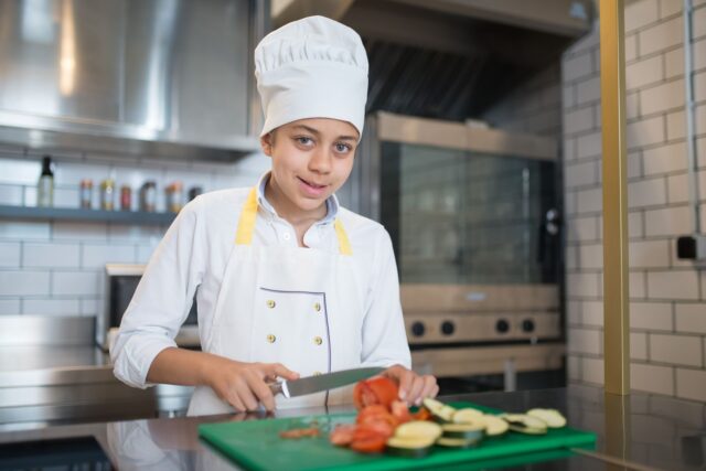 Bucătar care taie legume pe un blat din inox, într-o bucătărie profesională bine luminată